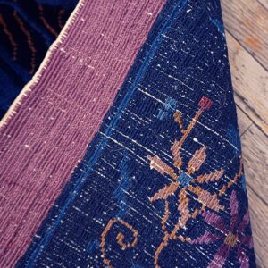 Close-up of a rug's folded backing, featuring mauve binding, dark blue, purple, and tan geometric patterns on wood.