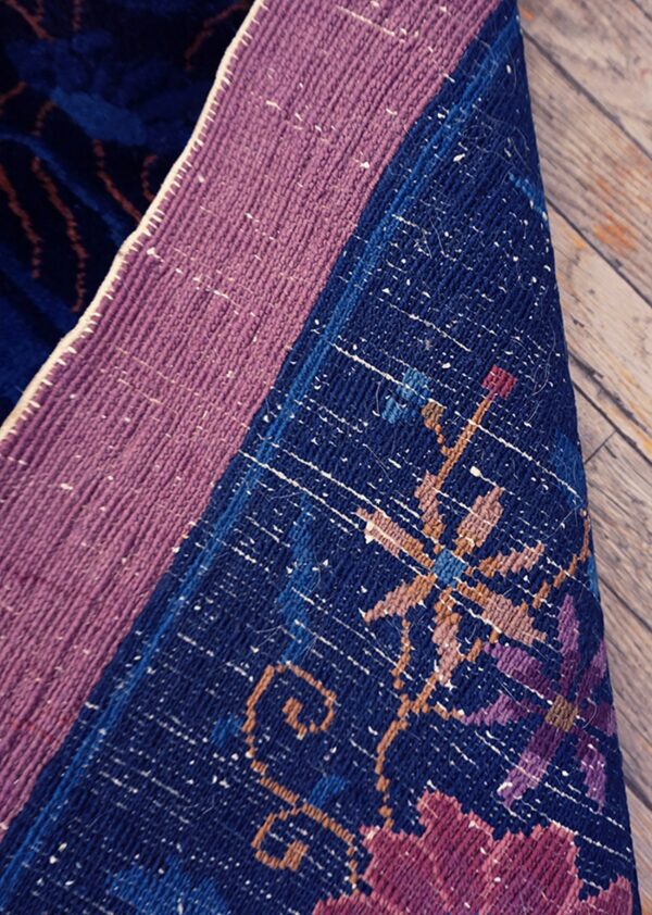 Close-up of a rug's folded backing, featuring mauve binding, dark blue, purple, and tan geometric patterns on wood.
