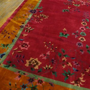 Red floral rug with orange and green borders rests on worn plank wood flooring.