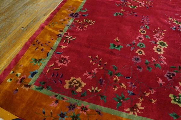 Red floral rug with orange and green borders rests on worn plank wood flooring.
