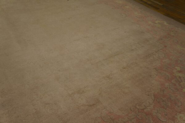 Low-pile beige rug field next to an ornate pink and yellow border on brown wooden flooring.