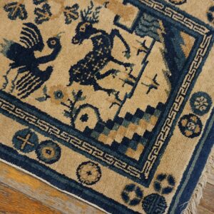 Tan rug with dark blue animal figures, patterned borders, and fringe rests on distressed wood flooring.