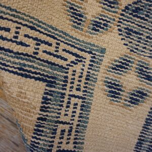 Textured, low-pile beige rug with dark blue geometric weave visible against distressed wood.