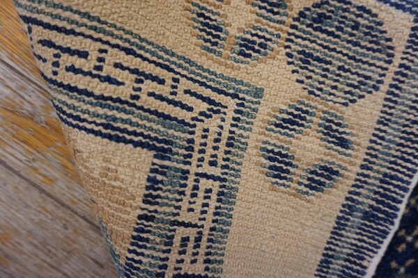 Textured, low-pile beige rug with dark blue geometric weave visible against distressed wood.
