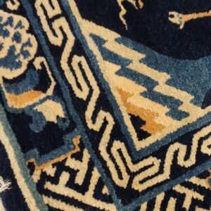 Detail of a navy blue rug with cream and teal geometric border patterns and visible fringe on wood flooring.