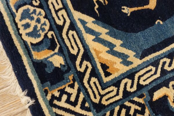 Detail of a navy blue rug with cream and teal geometric border patterns and visible fringe on wood flooring.