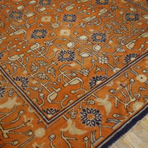 Angled view of an orange rug featuring intricate navy and tan botanical designs over rustic wood planks.