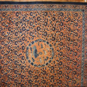 Orange rug with dark blue floral vines and borders, featuring a medallion, displayed on hardwood flooring.