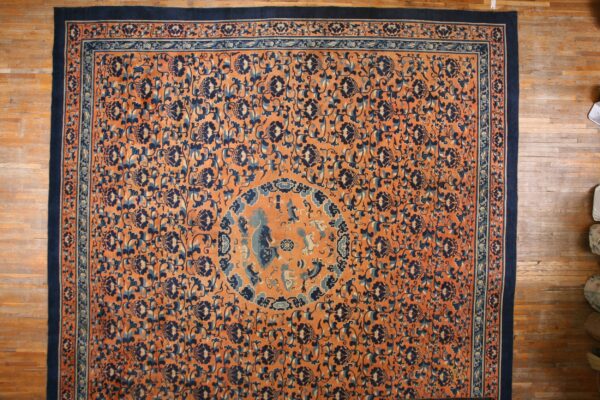 Orange rug with dark blue floral vines and borders, featuring a medallion, displayed on hardwood flooring.
