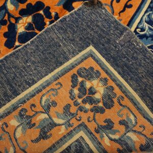Folded corner shows the orange, navy, and cream floral pile against the tightly woven blue rug backing.