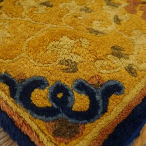 Textured rug corner in mustard yellow with dark blue scrollwork and layered borders on wood plank flooring.
