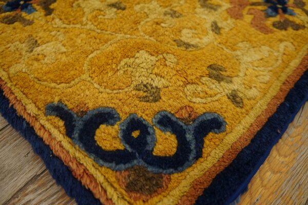 Textured rug corner in mustard yellow with dark blue scrollwork and layered borders on wood plank flooring.