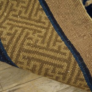 Curled rug corner shows tan woven texture, blue stripes, and the reverse side’s low pile geometric pattern.