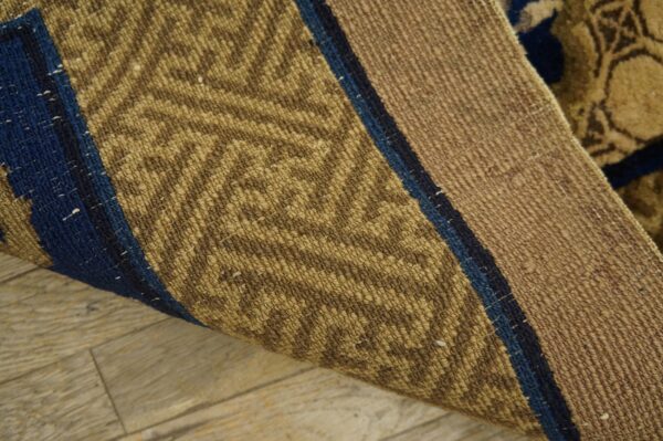 Curled rug corner shows tan woven texture, blue stripes, and the reverse side’s low pile geometric pattern.