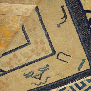 Beige rug with navy borders and stylized motifs is folded back on weathered yellow plank flooring.