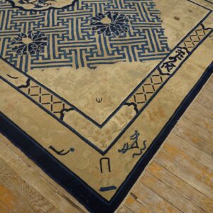 A beige rug featuring a dark blue geometric fretwork pattern and stylized floral motifs rests on worn wood flooring.