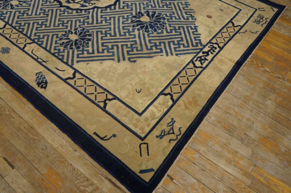 A beige rug featuring a dark blue geometric fretwork pattern and stylized floral motifs rests on worn wood flooring.