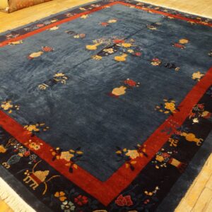 Large blue rug with scattered vase and floral patterns, featuring red and navy borders, on wood flooring.
