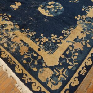 A worn navy blue rug with beige floral and vine patterns rests on rustic light brown plank flooring.