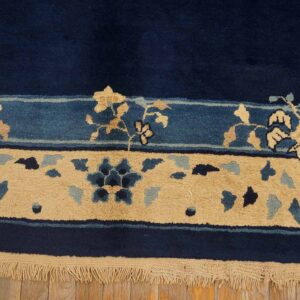 Navy rug with tan and blue floral borders rests on light wood planks showing visible fringe.
