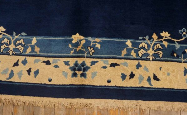 Navy rug with tan and blue floral borders rests on light wood planks showing visible fringe.