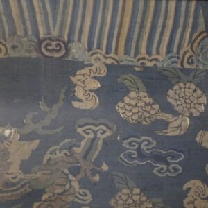 Faded blue and tan textile wall hanging in a gold frame displays mythical animals and floral patterns.