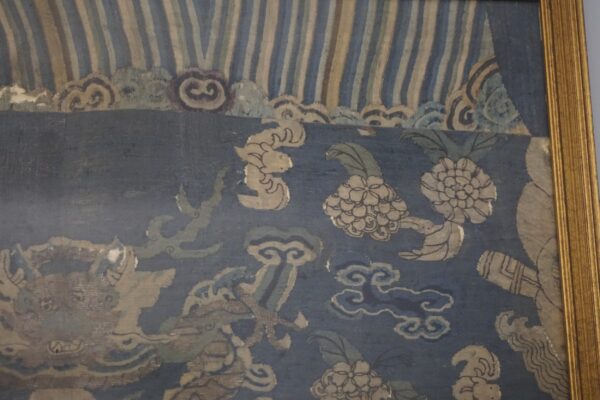 Faded blue and tan textile wall hanging in a gold frame displays mythical animals and floral patterns.