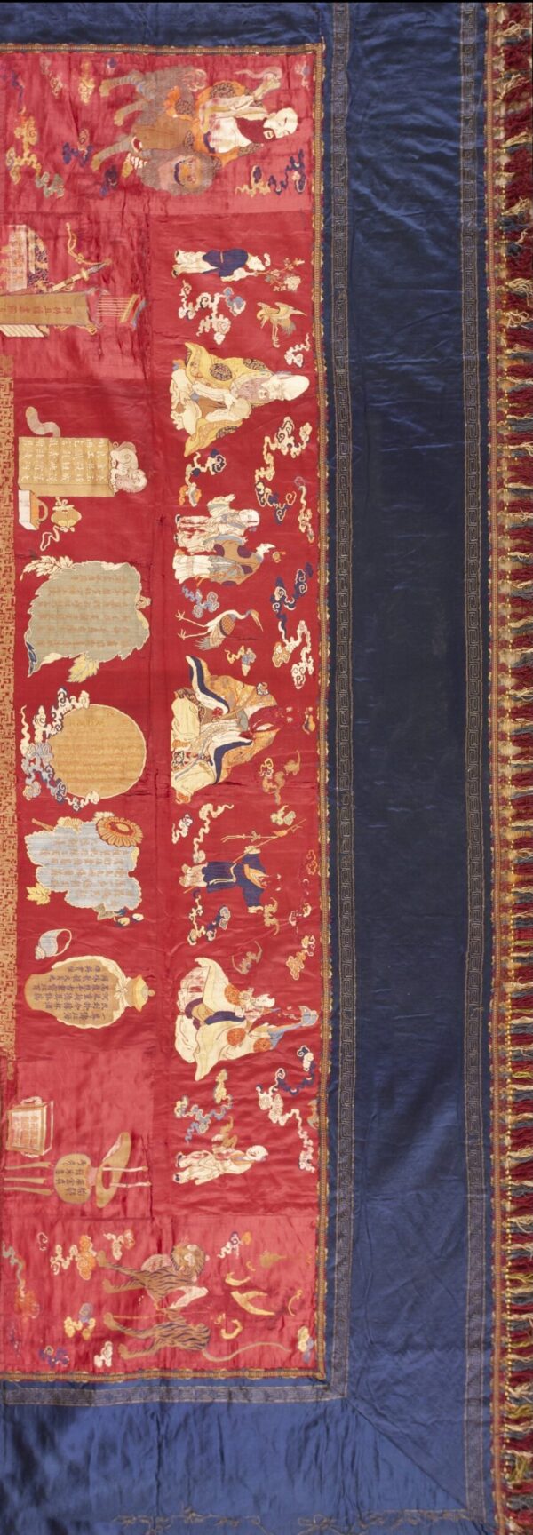 Shiny red textile featuring embroidered figures and script is bordered by blue fabric and elaborate fringe.