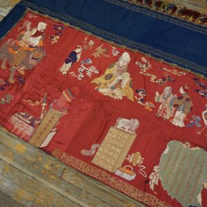 Detailed red embroidered textile with figural scenes and a blue border rests diagonally on distressed wooden planks.