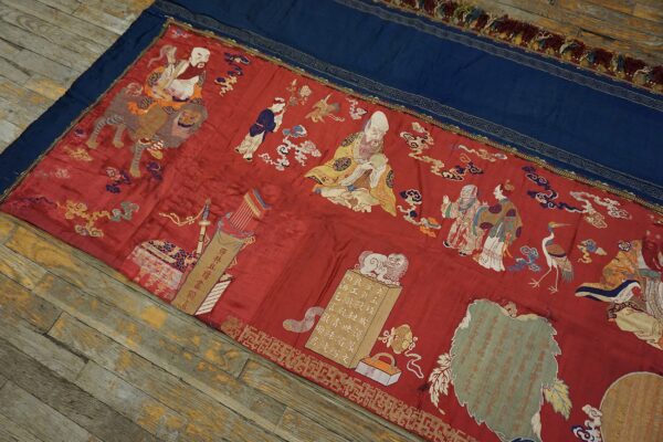 Detailed red embroidered textile with figural scenes and a blue border rests diagonally on distressed wooden planks.
