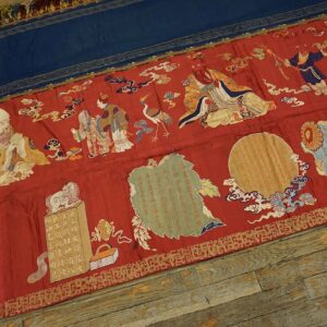 A flat red and blue textile banner with detailed figural embroidery lies on distressed wooden planks.