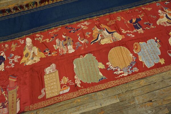 A flat red and blue textile banner with detailed figural embroidery lies on distressed wooden planks.