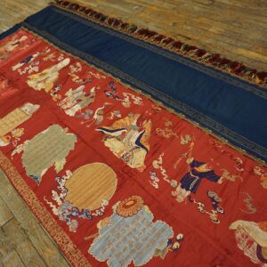 Long flatweave textile in red and blue with gold figural embroidery displayed on worn hardwood flooring.