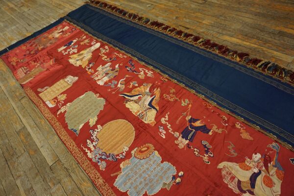 Long flatweave textile in red and blue with gold figural embroidery displayed on worn hardwood flooring.