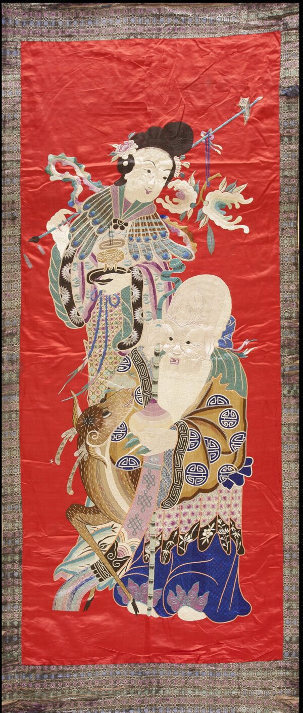 Red textile panel embroidered with two colorful figures and a deer, framed by a complex geometric border.