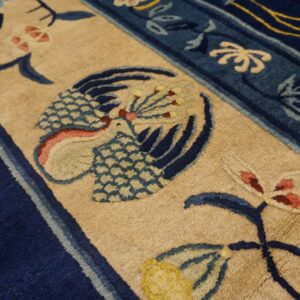 Thick-pile rug detail showing a beige field, deep blue border, and stylized bird and floral pattern.