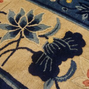 A close-up of a textured rug displays a tan background with dark and light blue botanical motifs.
