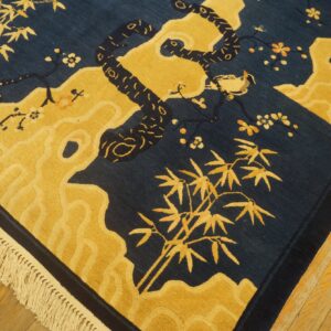 Close-up of a blue and gold rug featuring a snake, bird, bamboo, and carved fringe detail on wood floor.