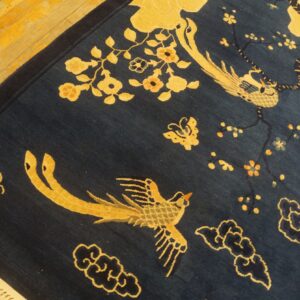 A dark blue rug with gold birds and flowers rests on light hardwood flooring, showing fringe.