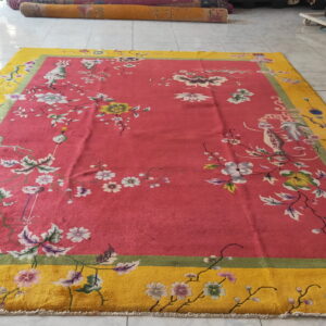 A red-pink rug with a wide yellow border featuring floral patterns lies on light tiled flooring.