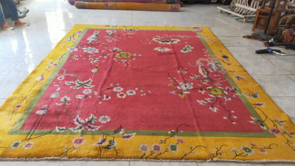 A red-pink rug with a wide yellow border featuring floral patterns lies on light tiled flooring.