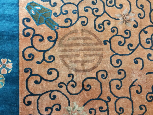 Close-up of a rug section featuring a peach field, teal border, dark scrolling patterns, and a worn central motif.