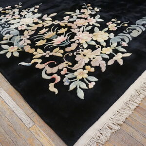 Black rug corner with pastel floral and scroll patterns on distressed light wood plank flooring.