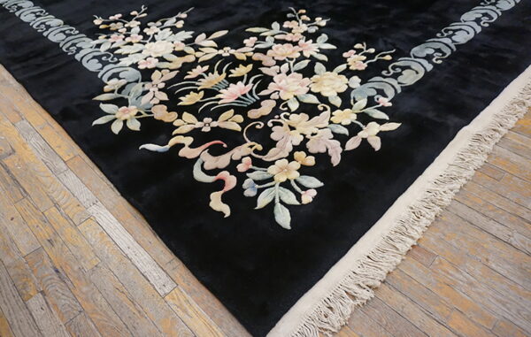Black rug corner with pastel floral and scroll patterns on distressed light wood plank flooring.