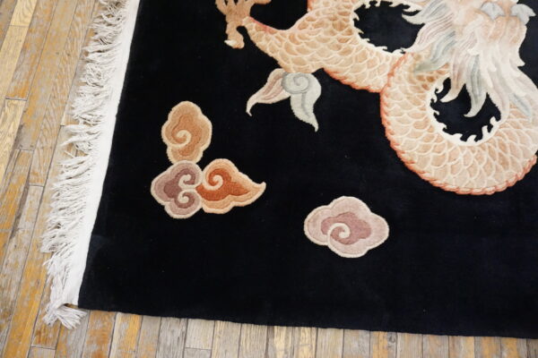Black rug with beige dragon and multicolored clouds sits on rustic wood planks, showing white fringe.