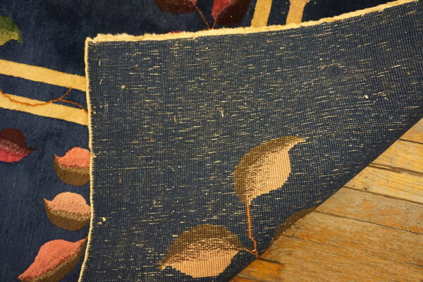 Navy floral rug corner lifted, displaying the low pile and woven backing over rustic wooden planks.