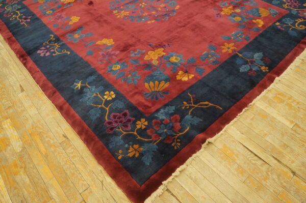 Maroon rug featuring a dark blue border and multicolored floral vase patterns on distressed wood flooring.