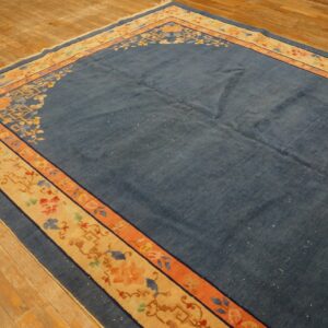 Blue rug with stylized floral and geometric beige border rests on worn wood flooring.