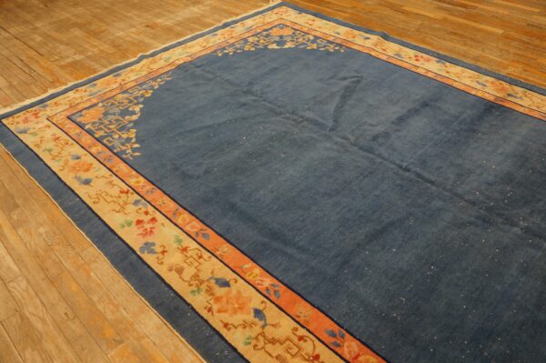 Blue rug with stylized floral and geometric beige border rests on worn wood flooring.