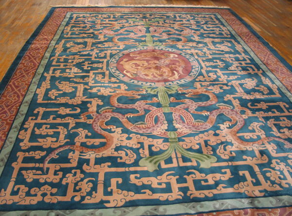 Teal rug featuring central dragon motifs and intricate tan geometric fretwork, displayed on wood plank flooring.